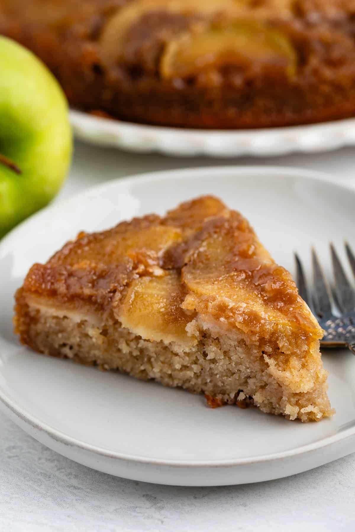 A slice of apple upside-down cake on a white plate with a fork beside it. A green apple and the rest of the cake are in the background.