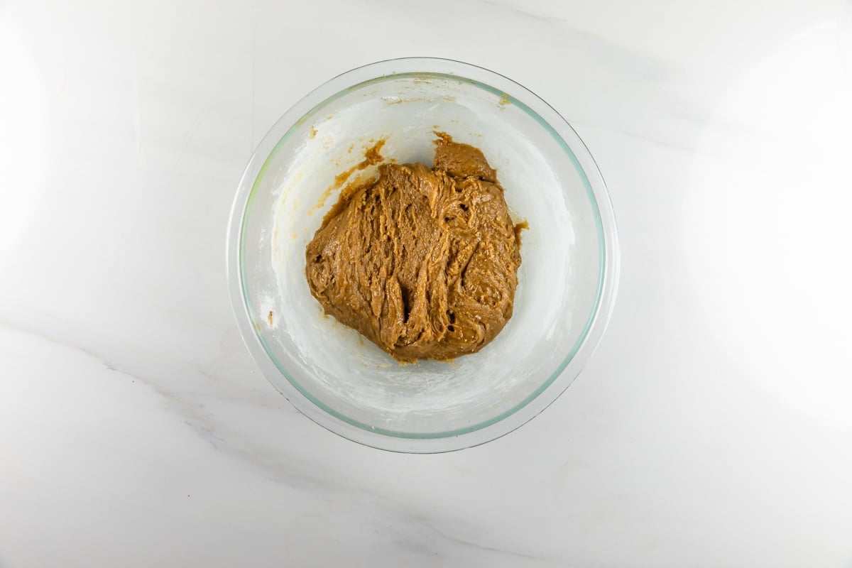 A glass bowl containing brown cookie dough sits on a white marble surface.