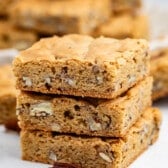 Three stacked blondie bars with visible pecan pieces sit on a white surface, with more blondies and scattered pecans in the background. The bars have a golden-brown color and a soft, chewy texture.