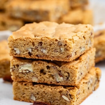 Three stacked blondie bars with visible pecan pieces sit on a white surface, with more blondies and scattered pecans in the background. The bars have a golden-brown color and a soft, chewy texture.