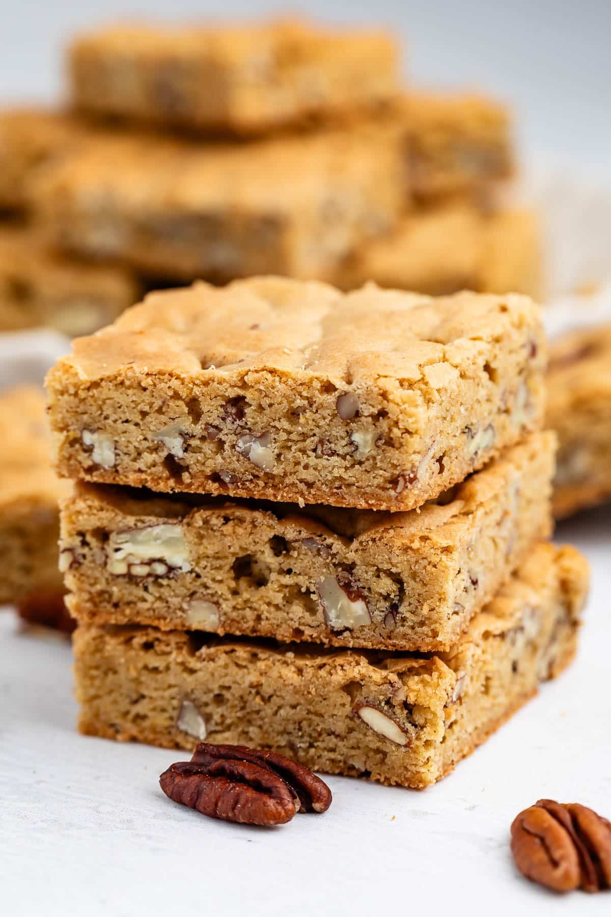 Three stacked blondie bars with visible pecan pieces sit on a white surface, with more blondies and scattered pecans in the background. The bars have a golden-brown color and a soft, chewy texture.