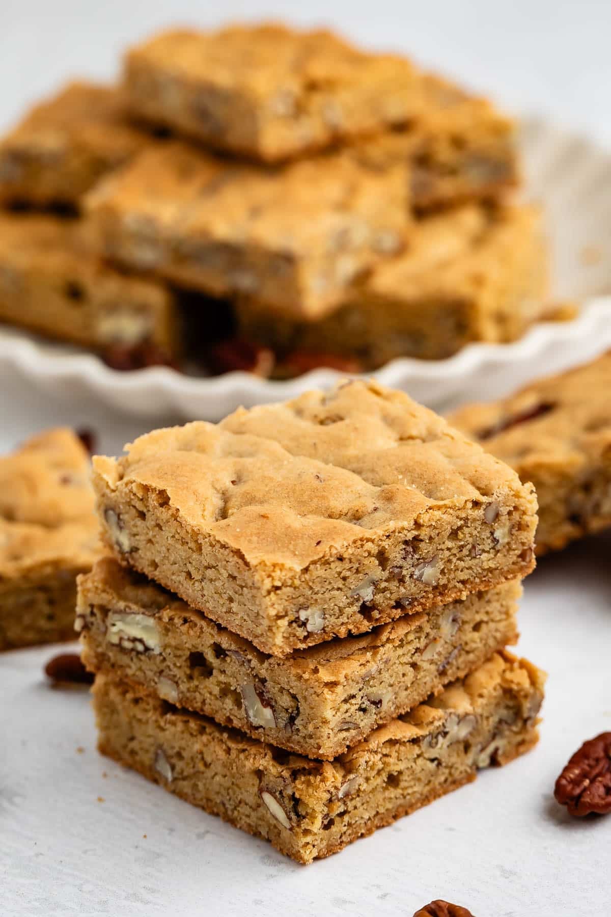 Three blondie bars with nuts are stacked in the foreground, while more bars are arranged on a plate in the background. Scattered pecans are visible on the white surface.