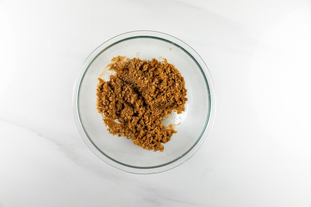 A clear glass bowl filled with light brown sugar on a white marble surface, viewed from above.