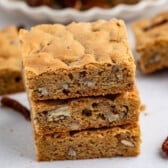 Three nutty blondie bars are stacked on a white surface, showing their golden-brown color and pieces of nuts inside. Loose pecans and more blondies are visible in the background.
