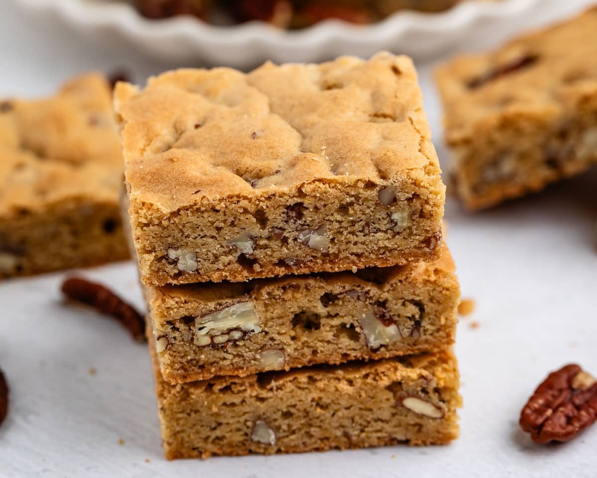 Three nutty blondie bars are stacked on a white surface, showing their golden-brown color and pieces of nuts inside. Loose pecans and more blondies are visible in the background.