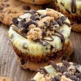 Mini cheesecakes topped with chocolate chips, cookie crumbles, and chocolate drizzle are displayed on a rustic wooden surface, surrounded by chocolate chip cookies.
