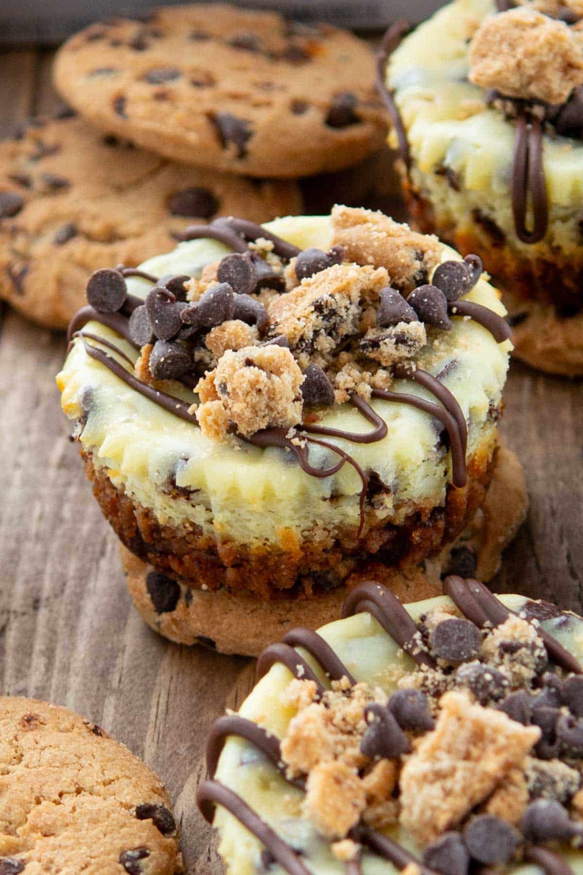 Mini cheesecakes topped with chocolate chips, cookie crumbles, and chocolate drizzle are displayed on a rustic wooden surface, surrounded by chocolate chip cookies.