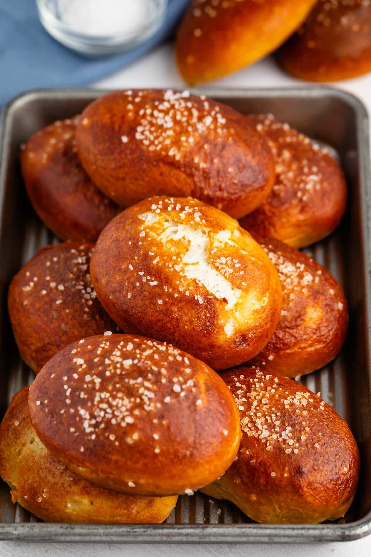 A tray filled with golden-brown pretzel rolls sprinkled with coarse salt, with a glass bowl of salt and more rolls visible in the background.