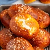 A tray of golden-brown pretzel rolls topped with coarse salt, with one roll showing a bite taken out of it.
