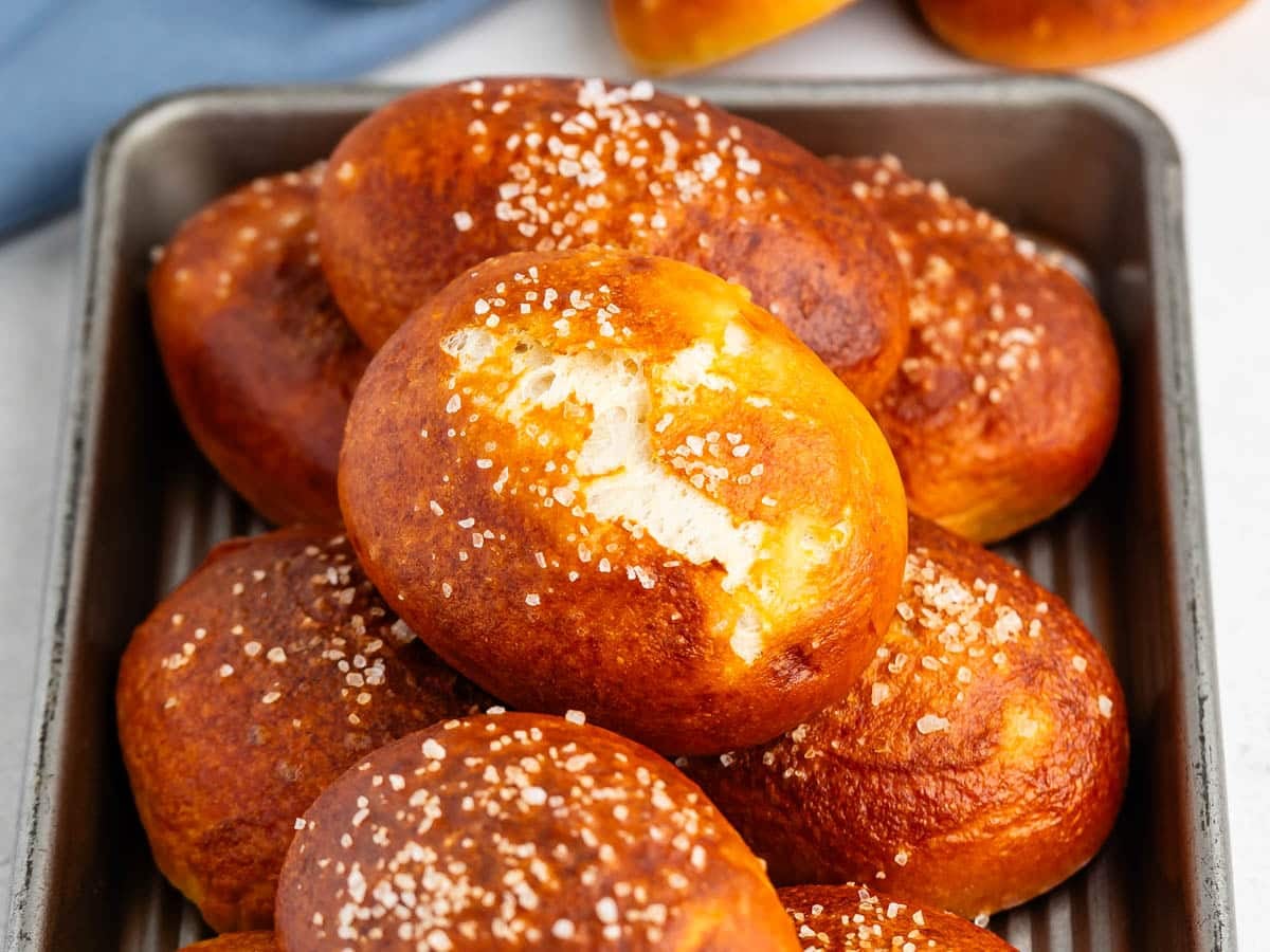 A tray of golden-brown pretzel rolls topped with coarse salt, with one roll showing a bite taken out of it.