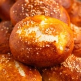 A close-up of golden-brown pretzel rolls topped with coarse salt, stacked on a baking tray.