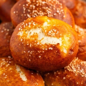 A close-up of golden-brown pretzel rolls topped with coarse salt, stacked on a baking tray.