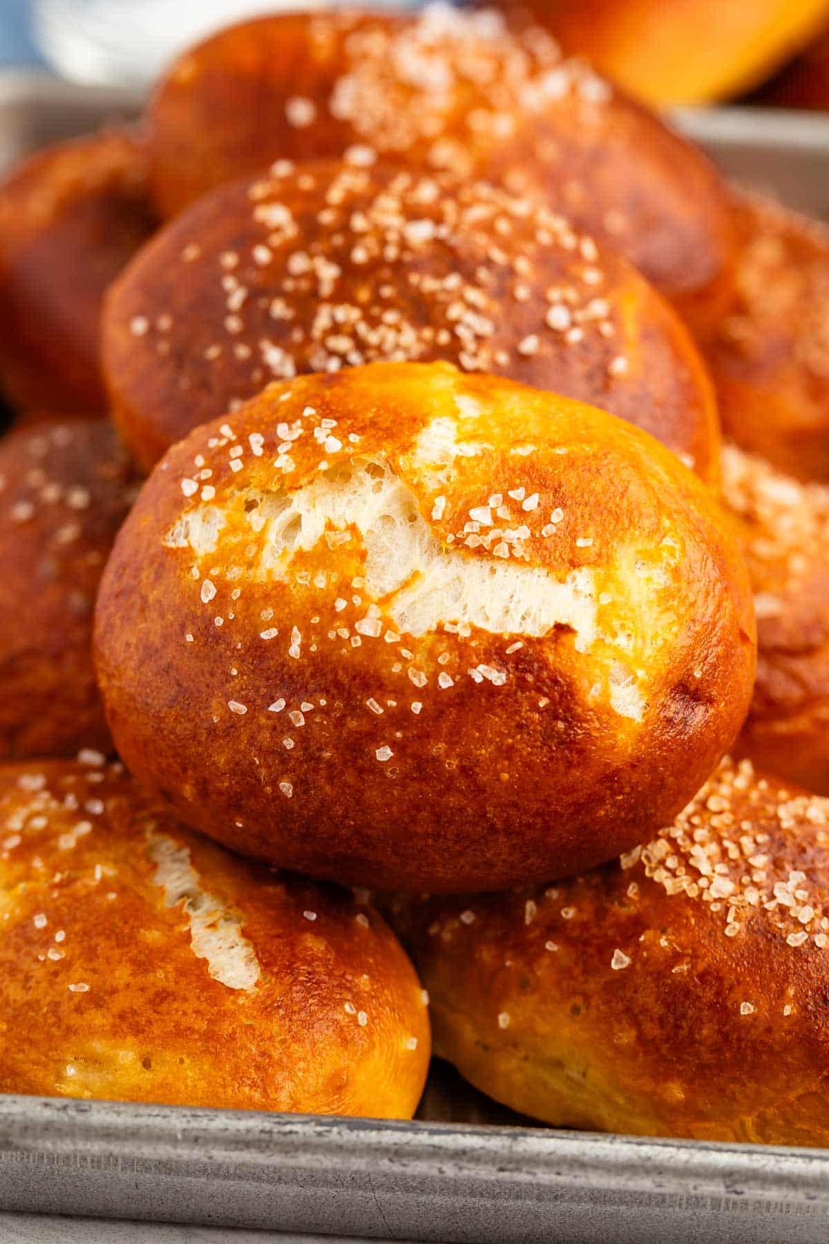 A close-up of golden-brown pretzel rolls topped with coarse salt, stacked on a baking tray.