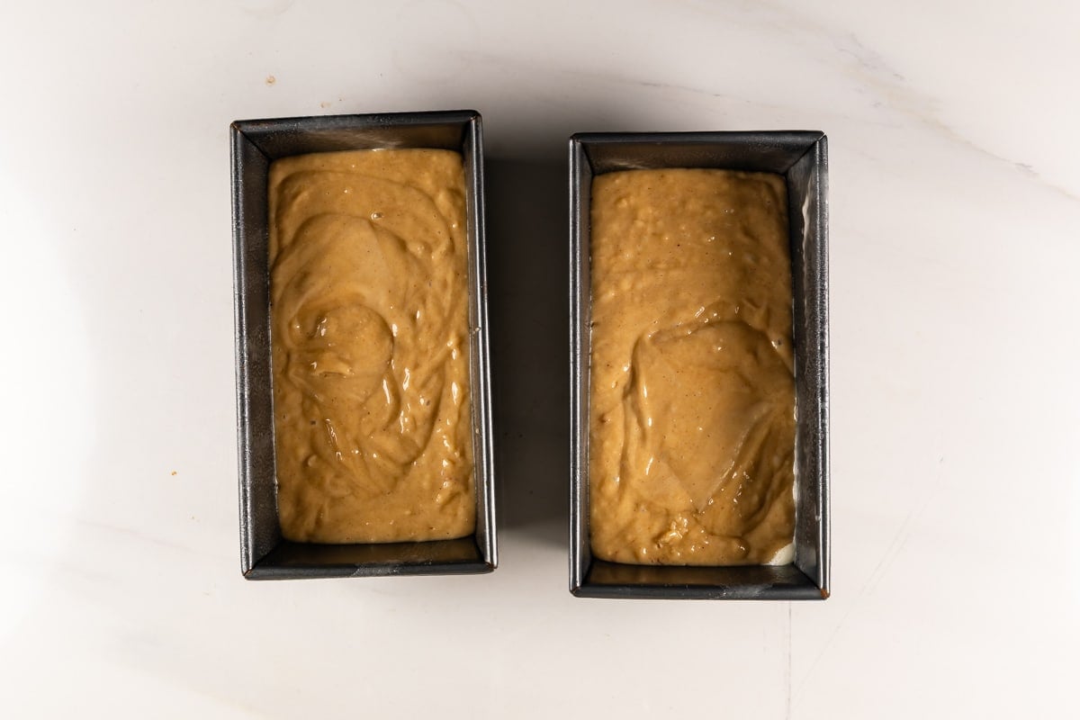 Two rectangular loaf pans filled with smooth, light brown batter sit side by side on a white countertop, ready to be baked.
