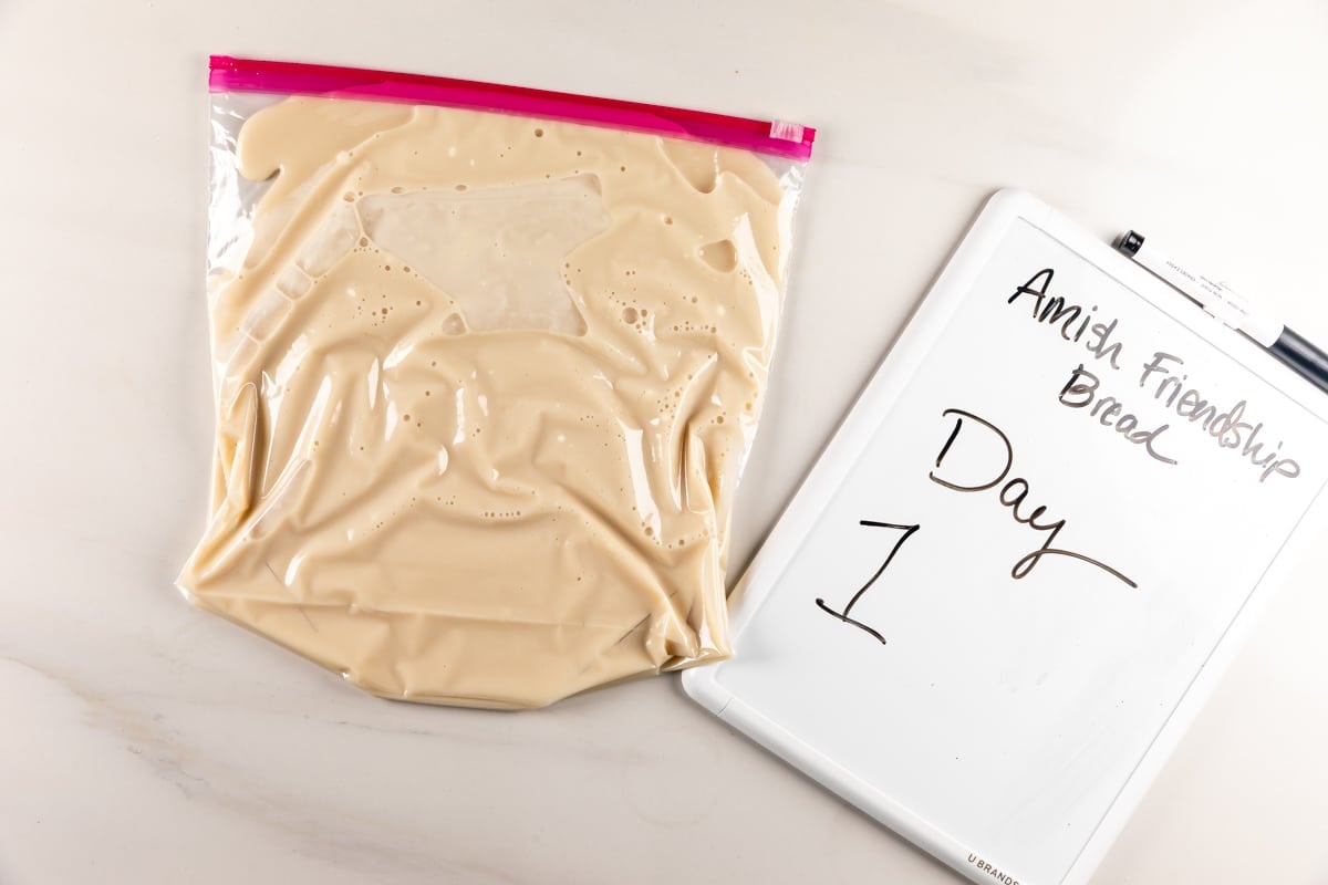 A plastic zip bag filled with beige batter sits next to a whiteboard that reads Amish Friendship Bread Day 1..