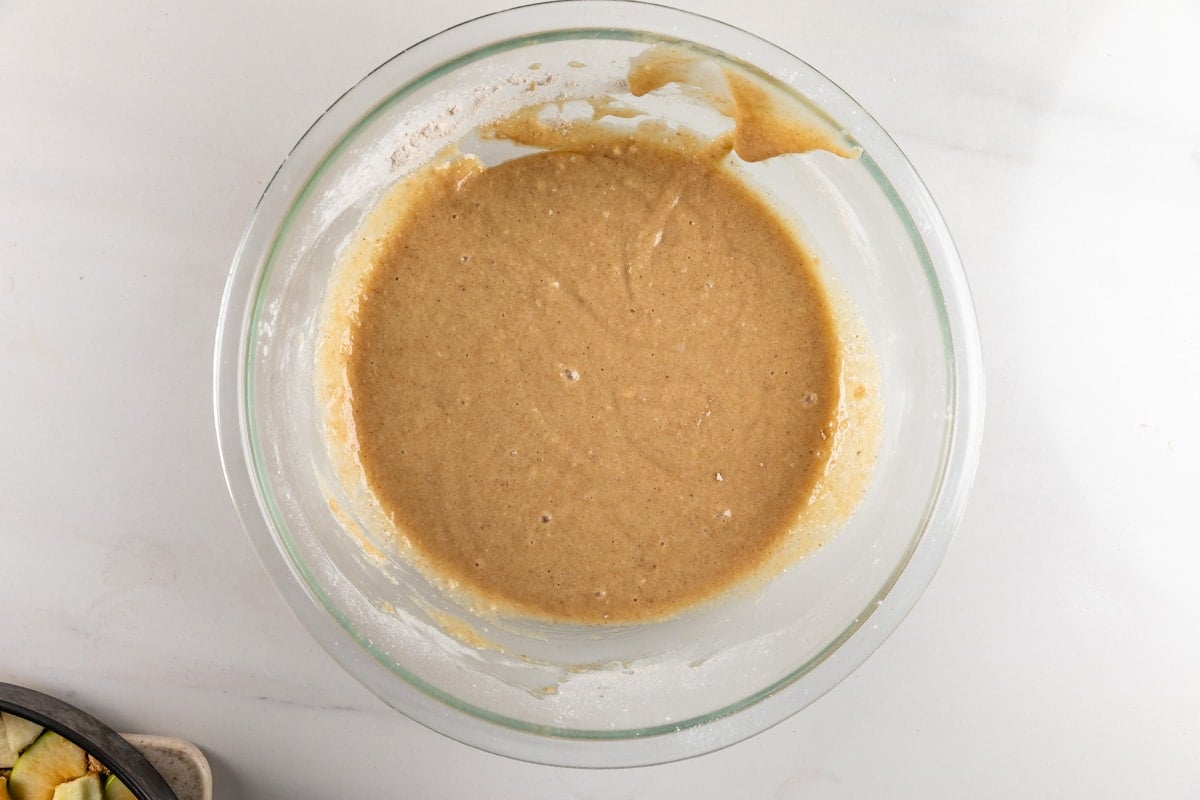 A glass bowl filled with light brown cake or muffin batter sits on a white countertop.