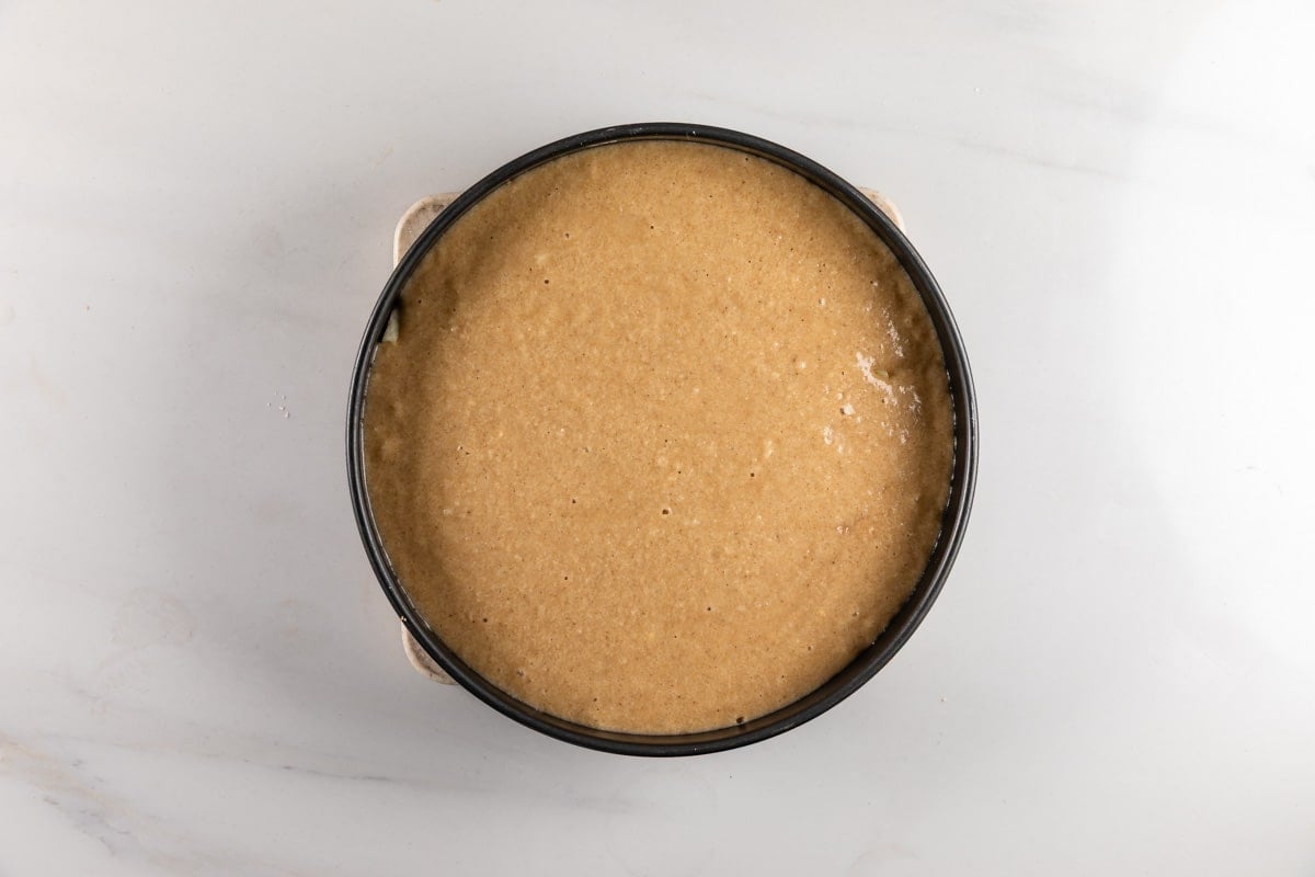 A round cake pan filled with light brown cake batter sits on a white marble surface, ready to be baked.