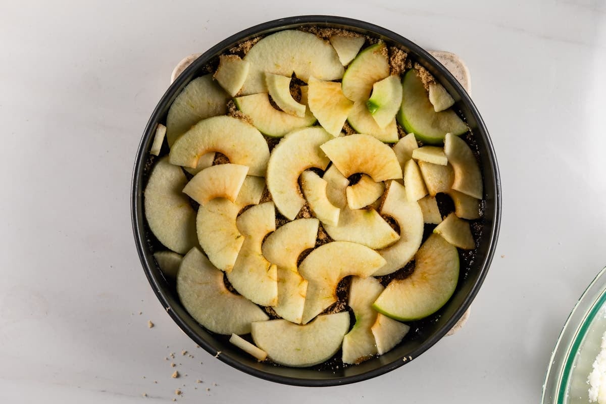 A round baking pan filled with sliced green apples arranged in layers on top of a brown crumbly mixture, sitting on a white surface.
