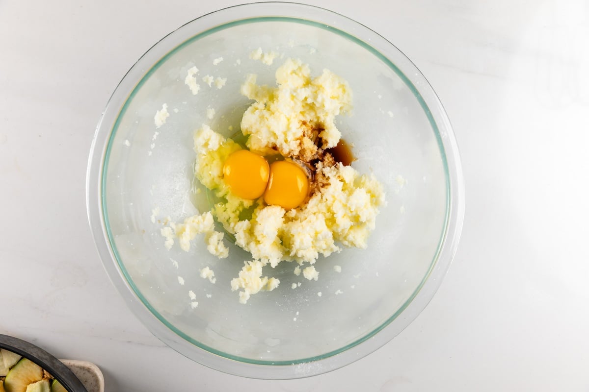 A glass bowl contains two raw eggs, creamed butter and sugar, and vanilla extract, ready for mixing, on a white countertop.
