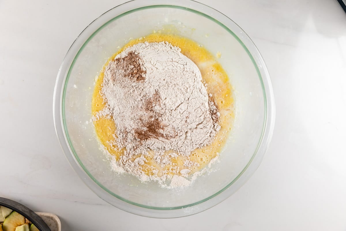 A glass bowl with flour, cinnamon, and wet ingredients being mixed on a white countertop, viewed from above.