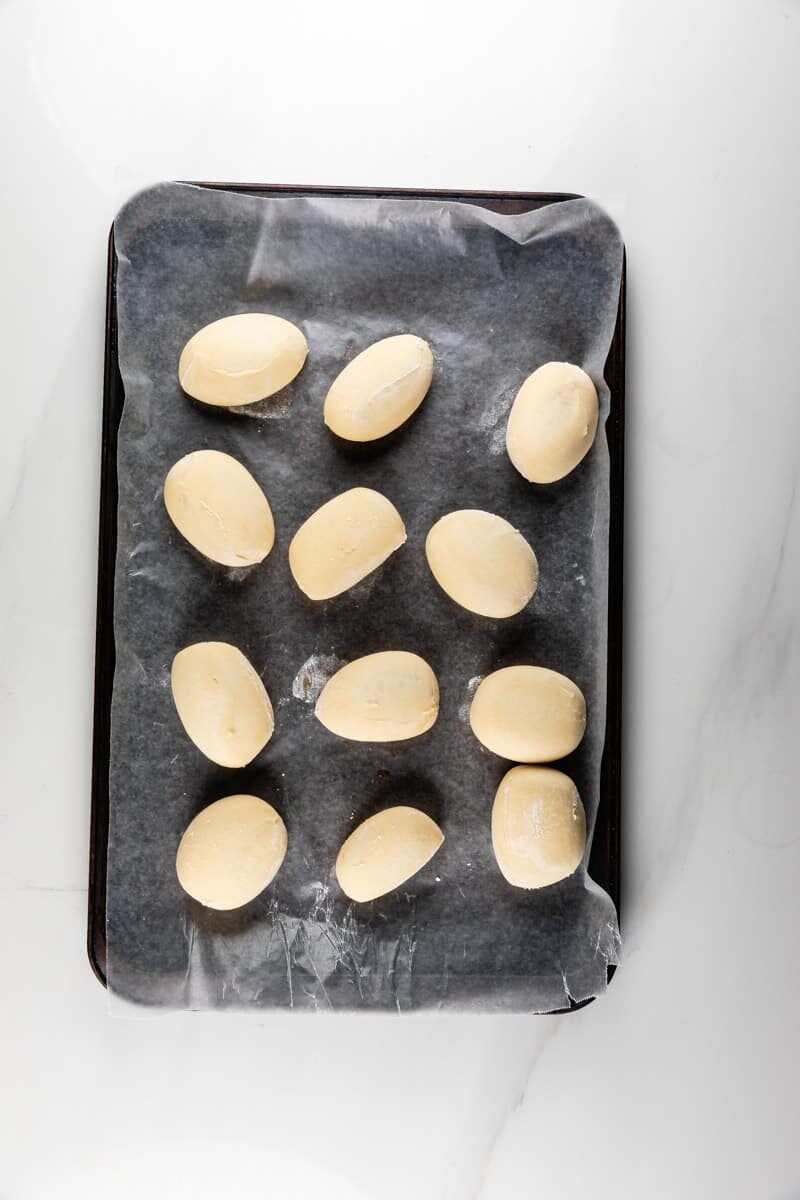 Twelve oval dough balls are arranged on a parchment-lined baking sheet, set on a white marble surface, ready to be baked.