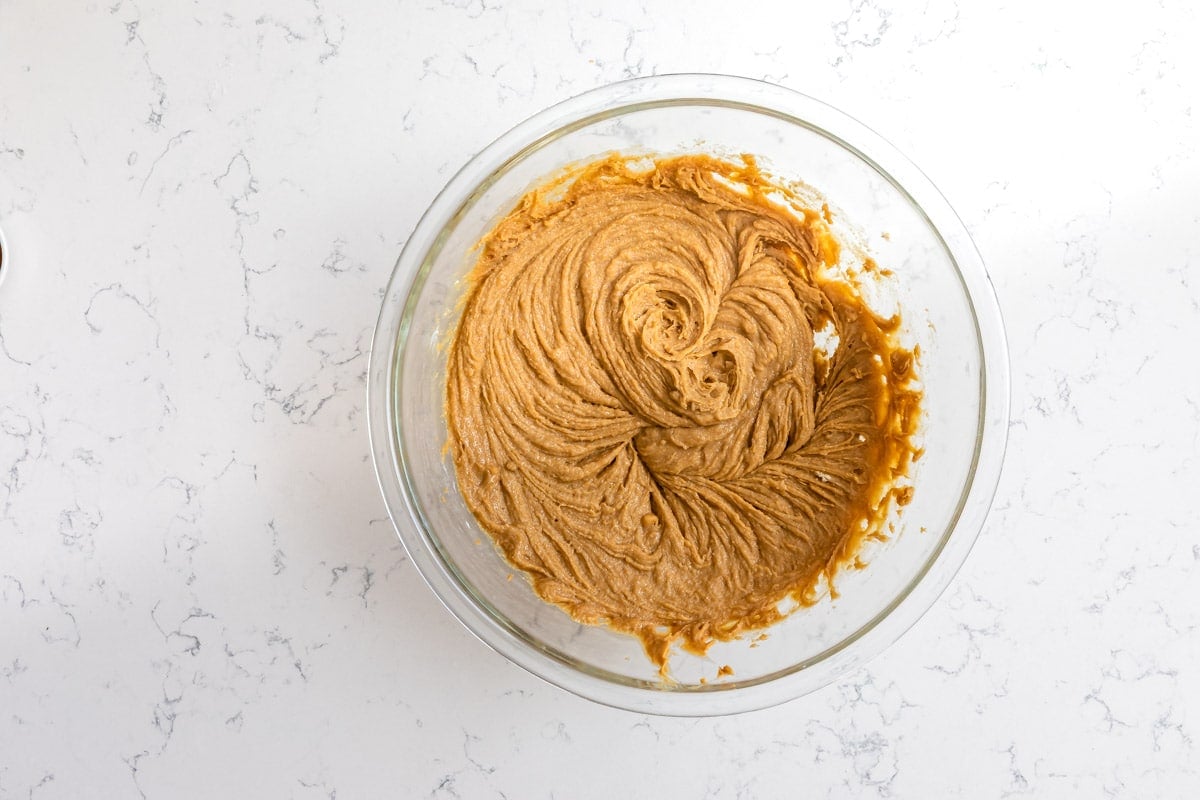 A glass bowl on a white marble countertop filled with light brown cookie dough batter, swirled smoothly in the center.