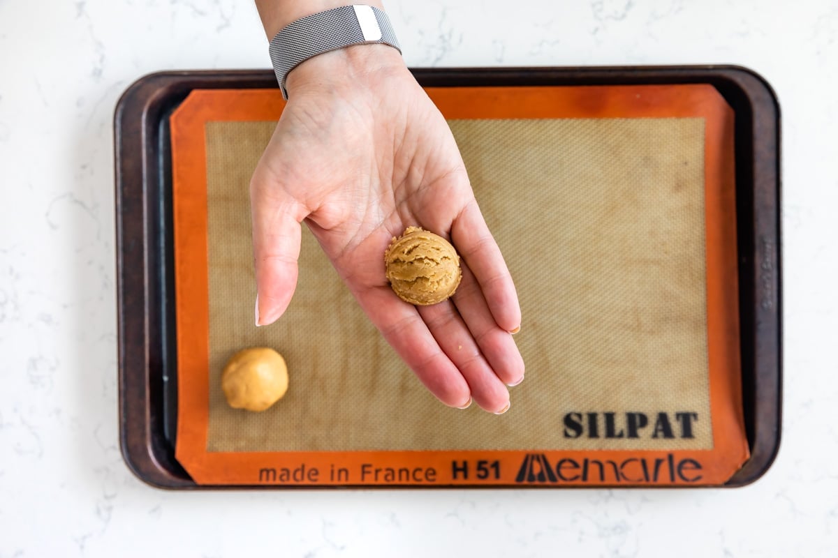 A hand holding a scoop of cookie dough above a Silpat-lined baking sheet, with another dough ball already placed on the baking sheet, ready for baking.