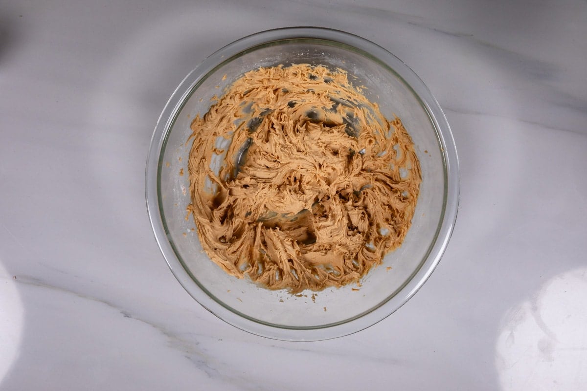A glass bowl filled with light brown cookie dough sits on a white marble surface. The dough appears to have been recently mixed, with swirled patterns visible.