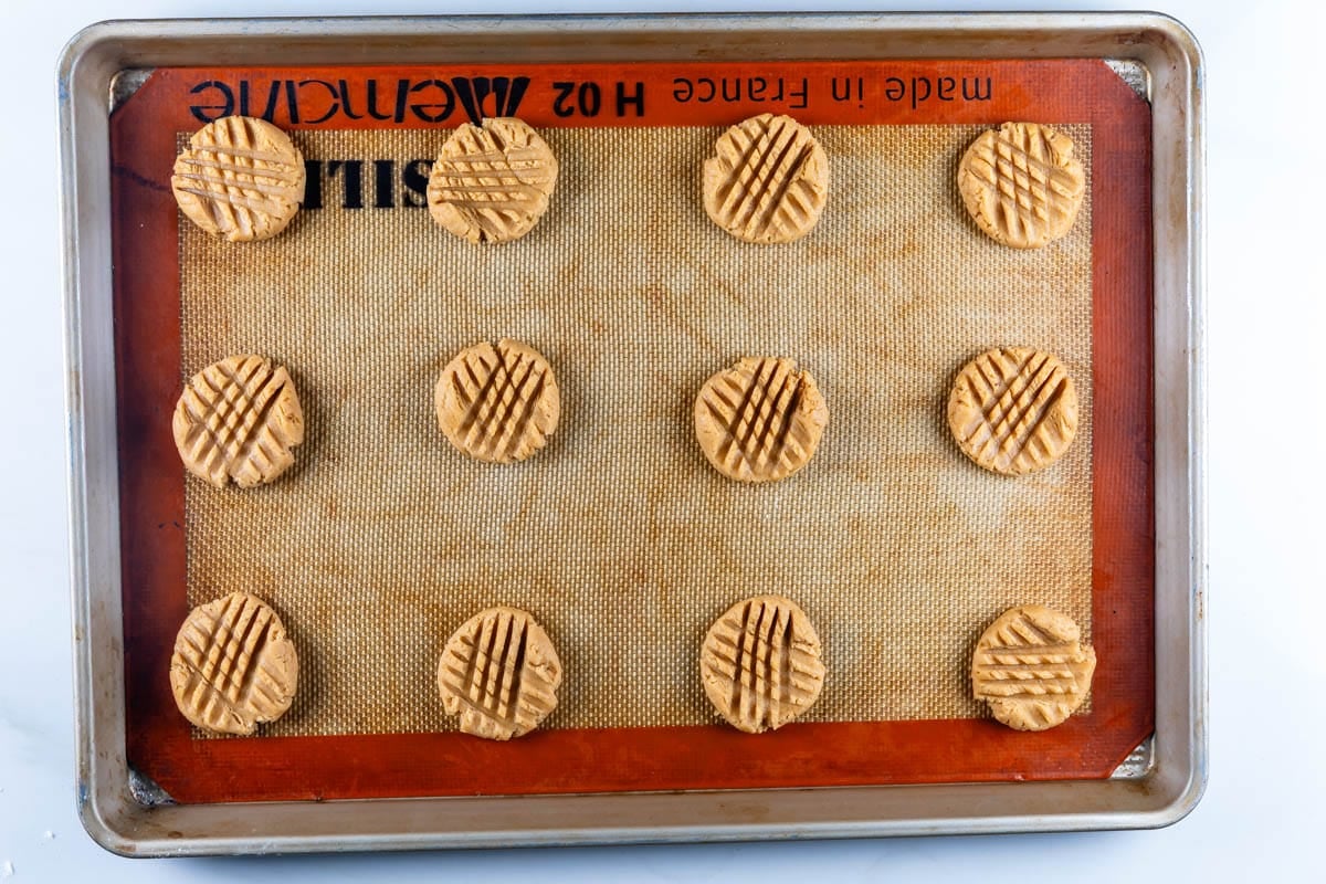 A baking sheet with 11 unbaked peanut butter cookies, evenly spaced on a silicone mat, each pressed with a crisscross pattern using a fork.