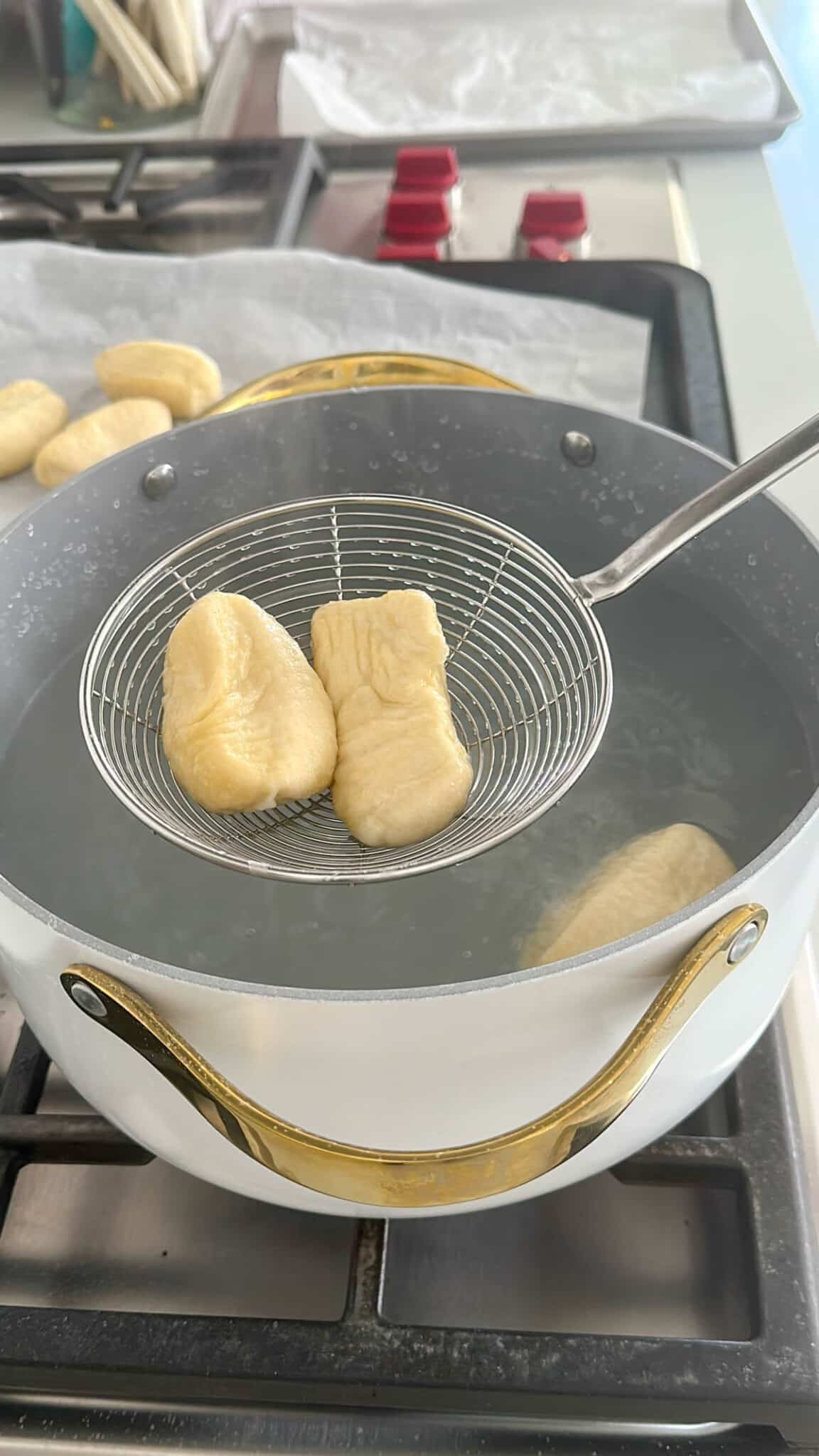 A wire skimmer lifts two pieces of dough from boiling water in a white pot with gold handles on a stove. More dough pieces rest on a tray in the background.