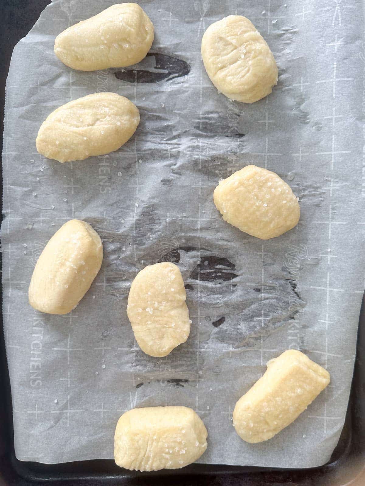 Eight pieces of raw gnocchi dough are arranged on parchment paper atop a baking tray, ready for baking. Some are lightly dusted with flour or salt.