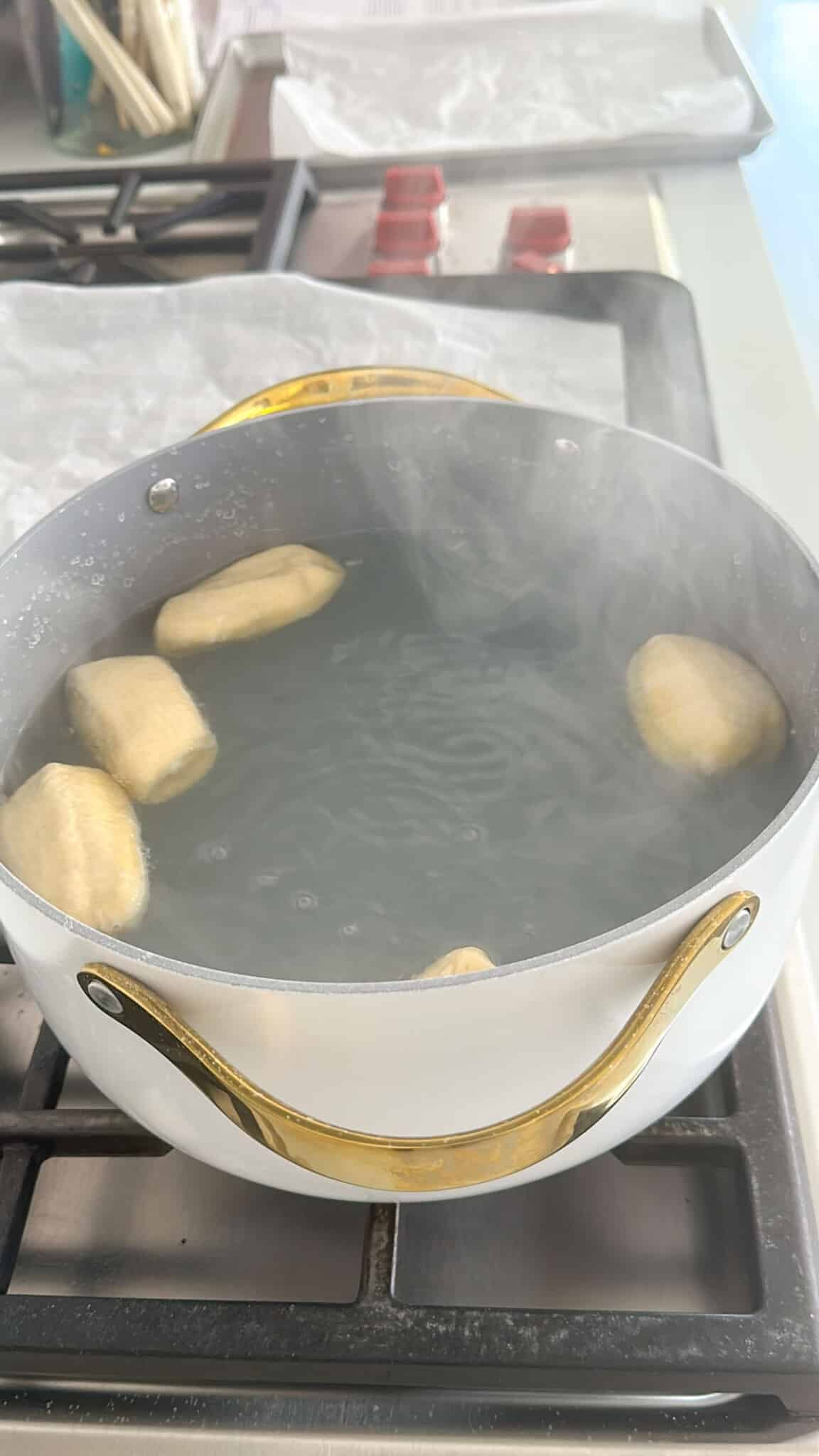 A white pot with gold handles is boiling six pieces of dough in water on a stovetop. Steam is rising, and a baking tray with parchment paper is visible in the background.