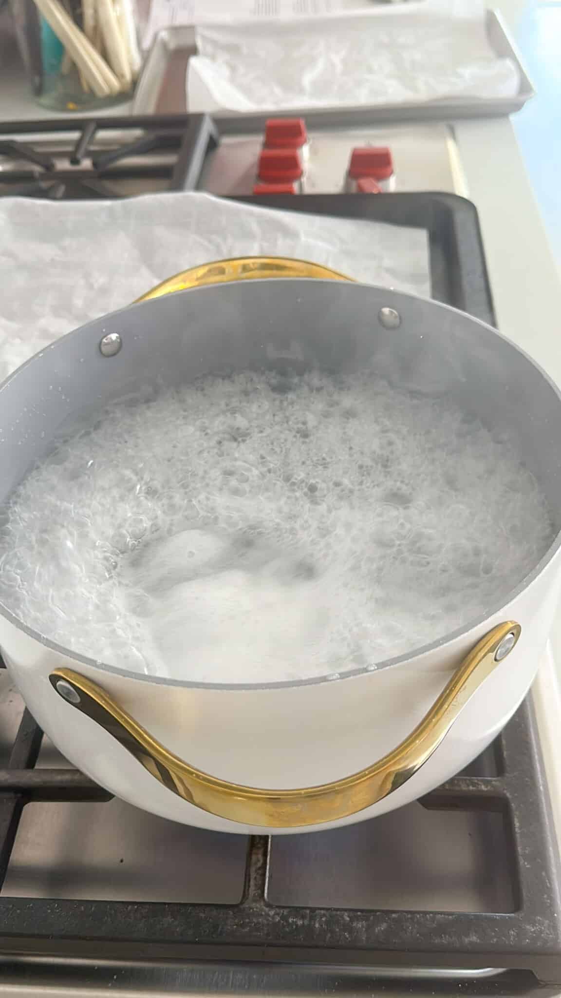 A silver pot with gold handles filled with boiling water sits on a stove, with steam rising and bubbles forming at the surface.