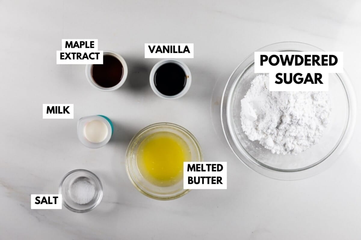 Six labeled ingredients for a recipe are displayed on a white surface: maple extract, vanilla, milk, melted butter, powdered sugar in a bowl, and salt in a small dish.