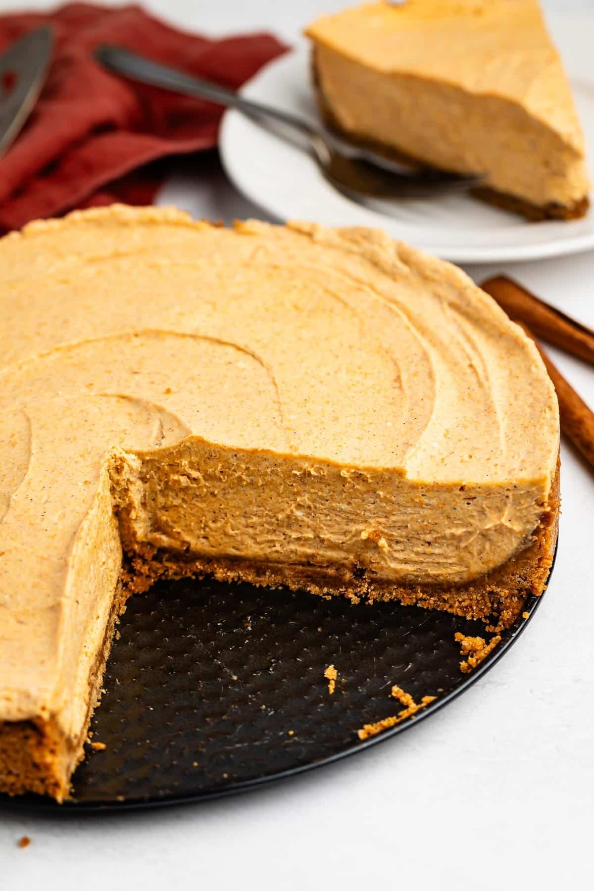 A pumpkin cheesecake with a graham cracker crust sits on a black plate, missing one slice. A single slice is served on a white plate in the background.