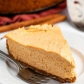 A slice of creamy pumpkin cheesecake sits on a white plate with a fork beside it. The background shows the rest of the cheesecake and a pie server.