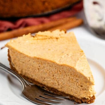 A slice of creamy pumpkin cheesecake sits on a white plate with a fork beside it. The background shows the rest of the cheesecake and a pie server.