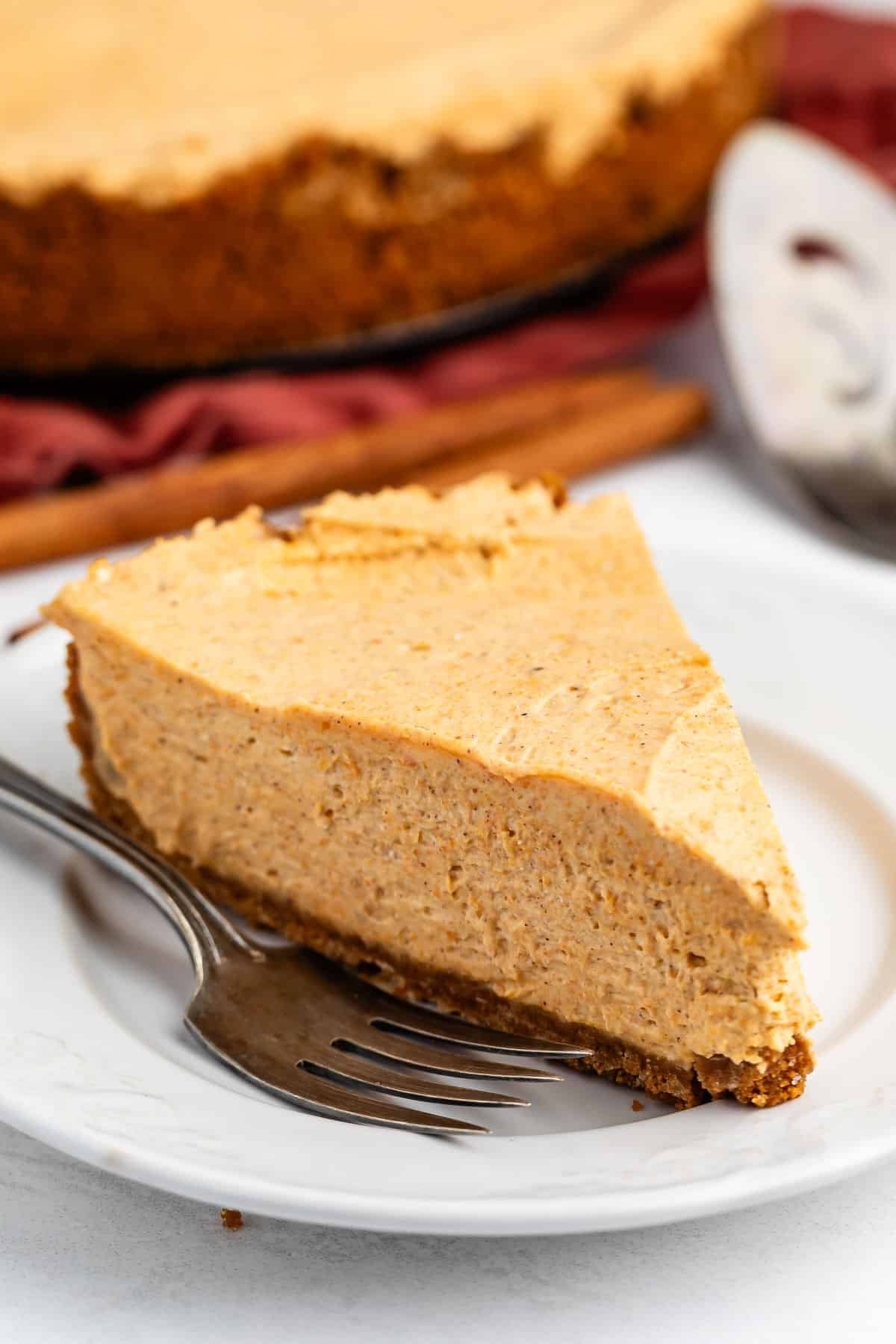 A slice of creamy pumpkin cheesecake sits on a white plate with a fork beside it. The background shows the rest of the cheesecake and a pie server.