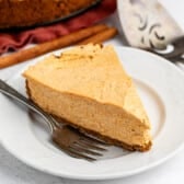 A slice of creamy pumpkin cheesecake sits on a white plate with a fork beside it. A cinnamon stick and a pie server are visible in the background.