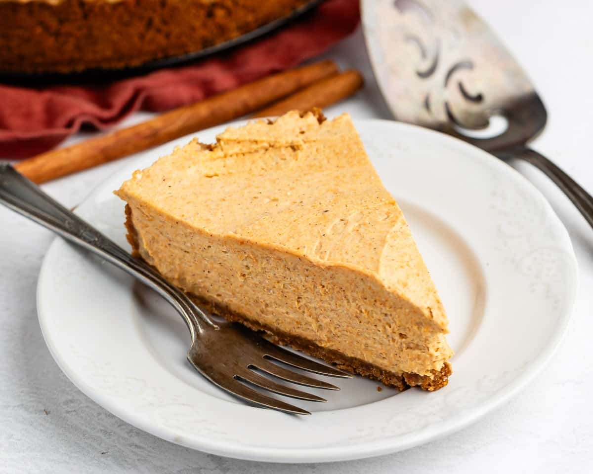 A slice of creamy pumpkin cheesecake sits on a white plate with a fork beside it. A cinnamon stick and a pie server are visible in the background.