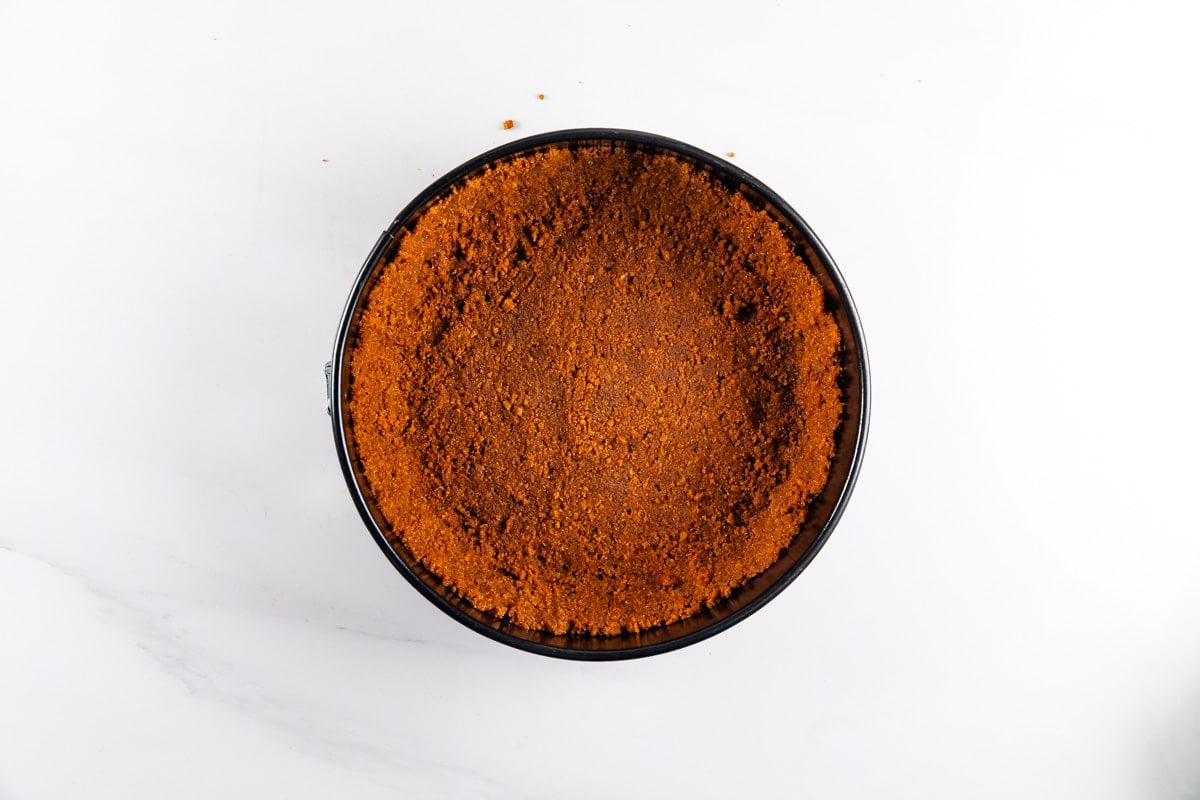 A round pan filled with a pressed, crumbly brown graham cracker crust, viewed from above on a white surface.
