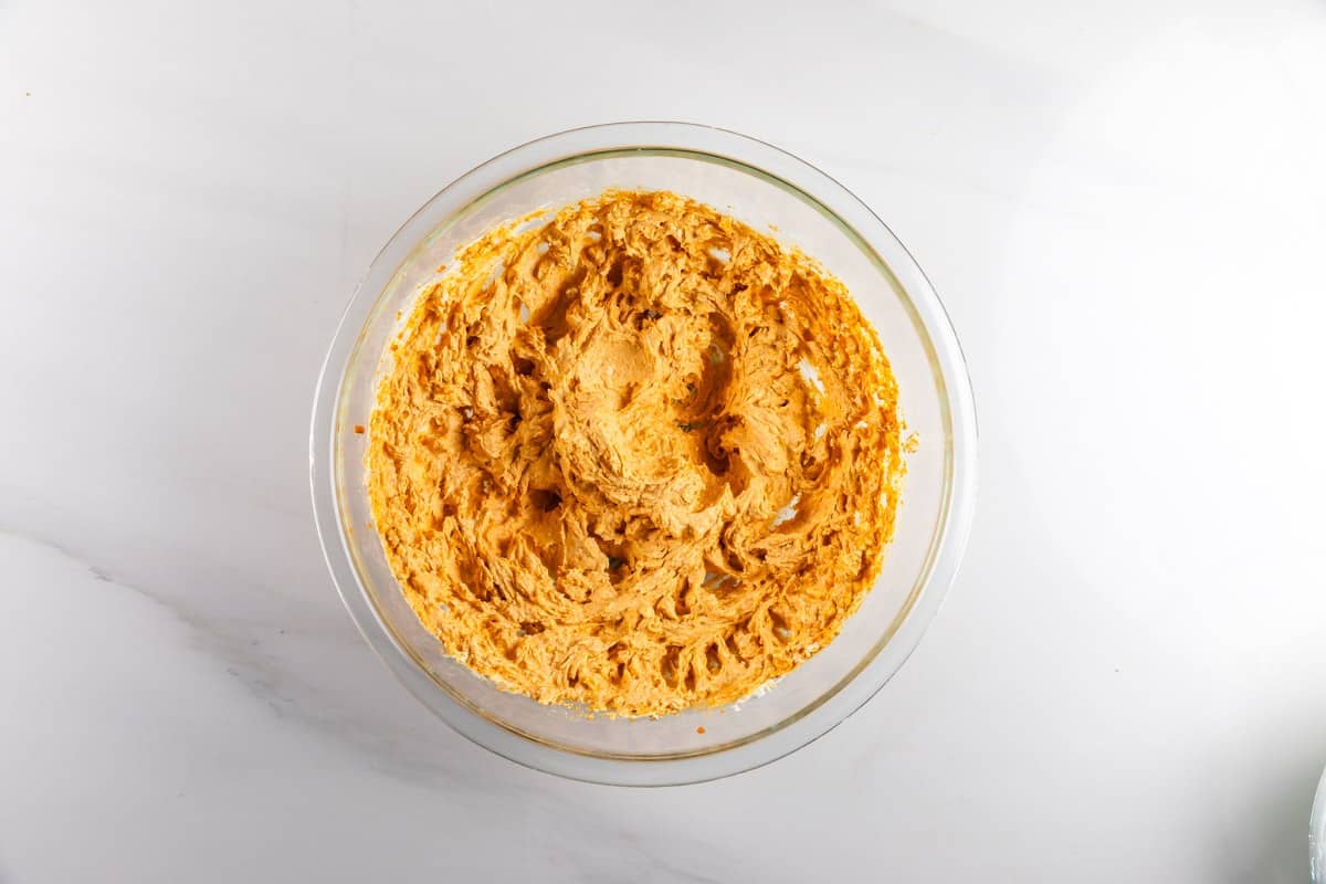 A glass bowl filled with a creamy, orange mixture sits on a white countertop. The mixture appears smooth with textured swirls on top.
