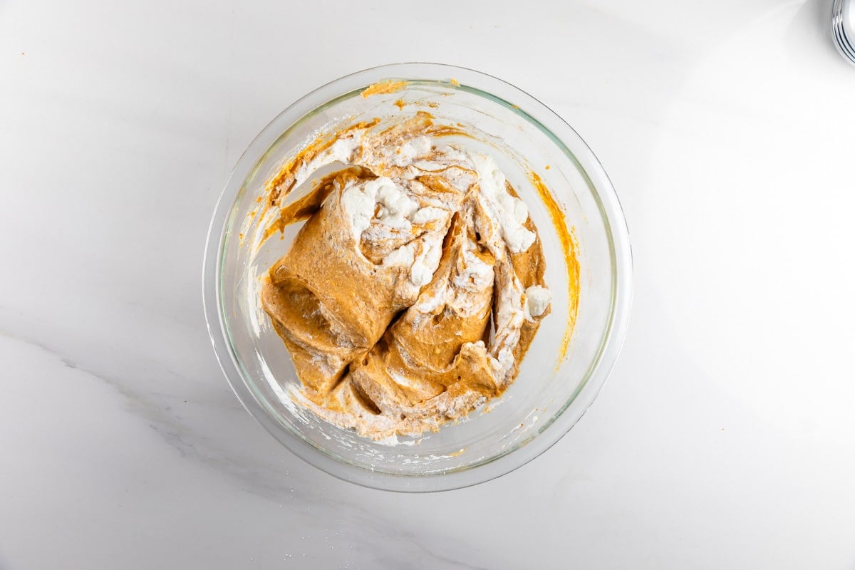 A glass bowl on a white surface holds partially mixed dough with visible flour and a brown batter, suggesting the beginning stages of baking.