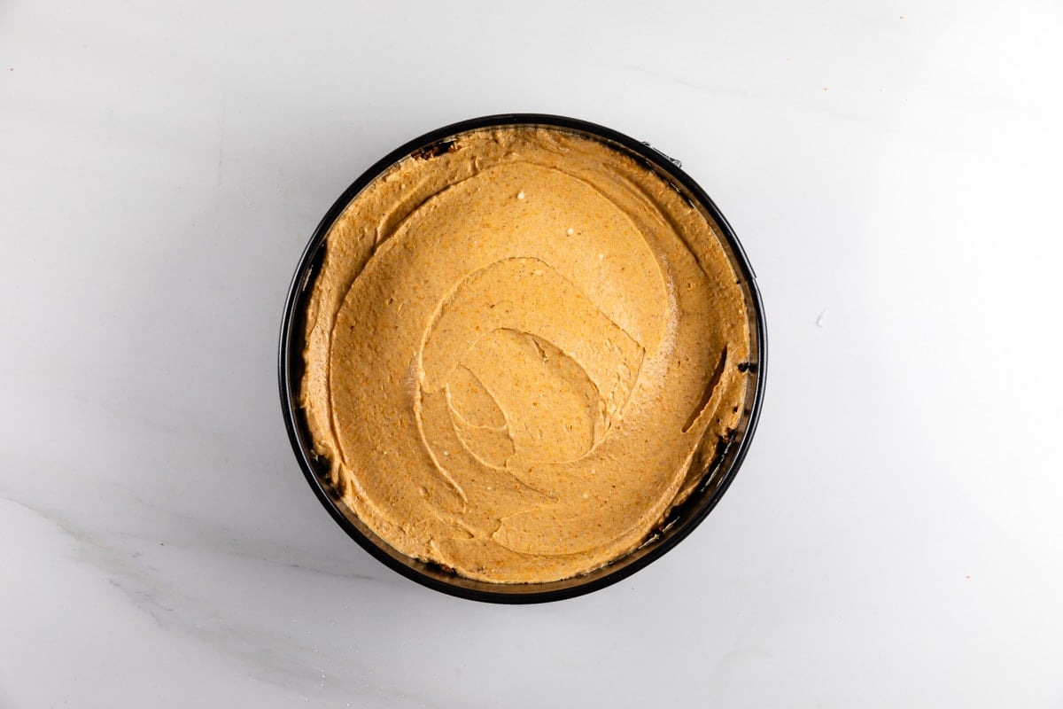 A round pan filled with a smooth, light brown creamy mixture, likely a cheesecake filling, sits on a white surface.