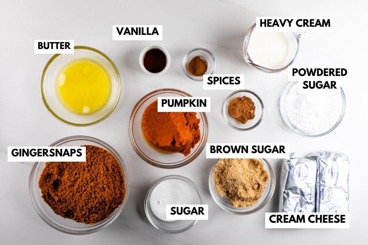 Overhead view of labeled baking ingredients in bowls on a white surface: butter, vanilla, pumpkin, spices, heavy cream, powdered sugar, brown sugar, cream cheese, sugar, and gingersnaps.