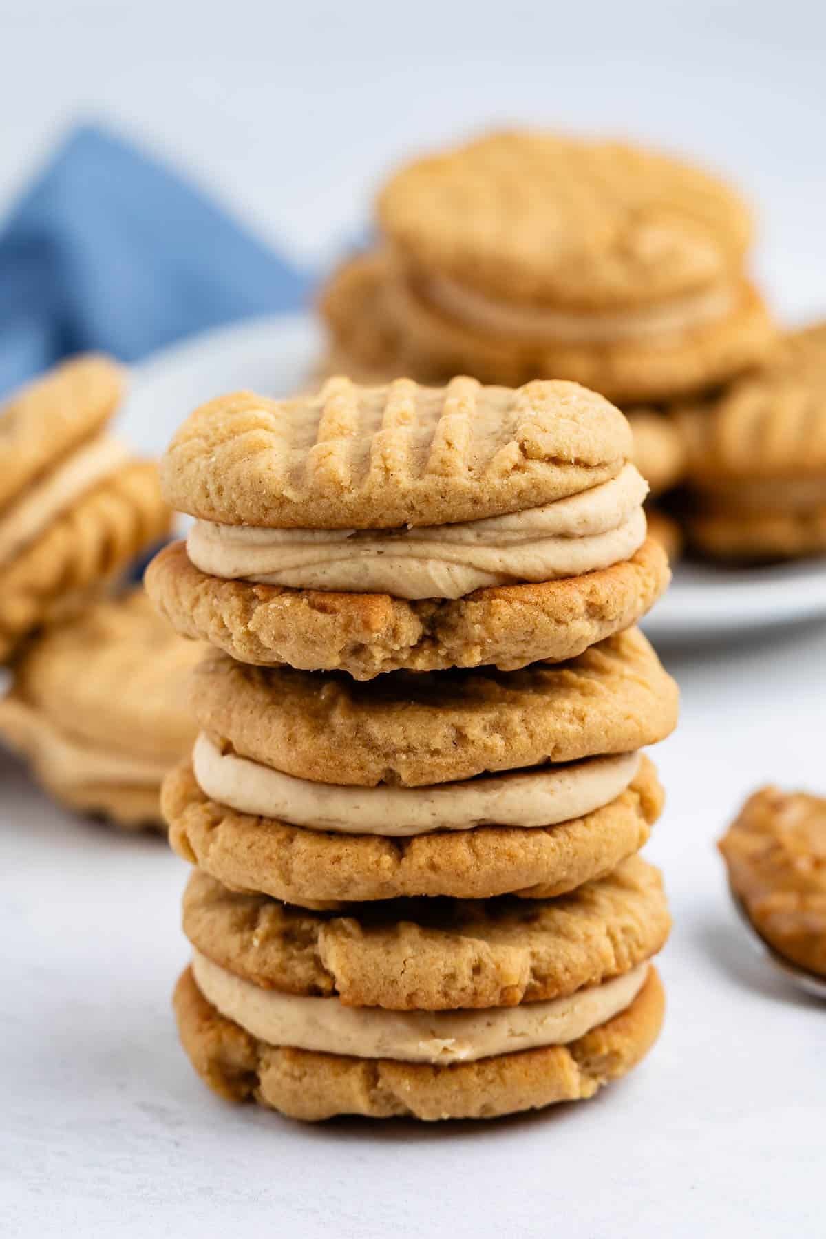 A stack of four peanut butter sandwich cookies filled with creamy peanut butter filling, with more cookies and a blue napkin blurred in the background.