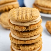 A stack of peanut butter sandwich cookies with a creamy filling, set on a white surface. More cookies and a blue cloth are visible in the background.