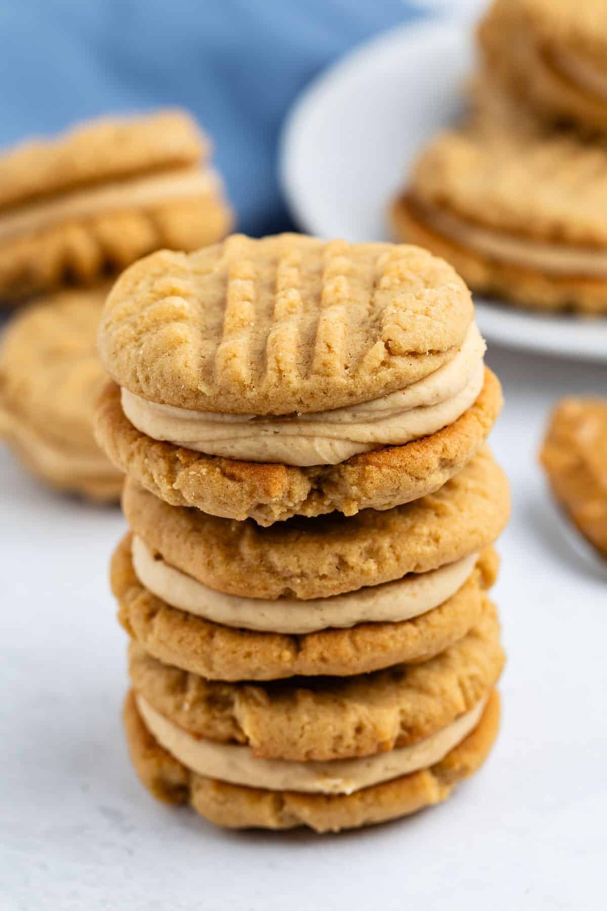 A stack of peanut butter sandwich cookies with a creamy filling, set on a white surface. More cookies and a blue cloth are visible in the background.