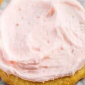 A close-up of a sugar cookie topped with a thick layer of light pink frosting, resting on a piece of parchment paper.
