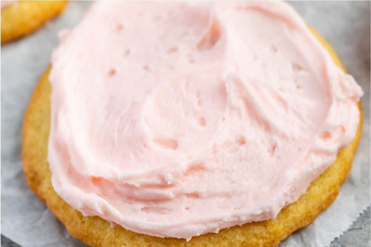 A close-up of a sugar cookie topped with a thick layer of light pink frosting, resting on a piece of parchment paper.